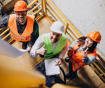 Three Happy Construction Workers In Uniforms And Hard Hats Working Outdoors On Scissor Lift And Giving Thumbs Up