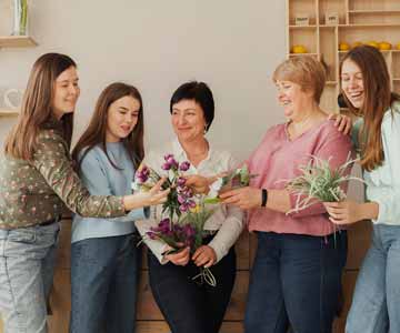 Five Happy Women From Different Generations In Bright Living Room Gathered Around Bouquet Of Purple Flowers
