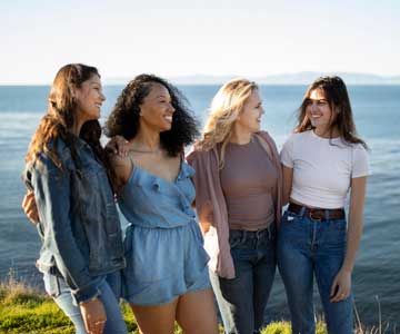 Four Smiling Young Women Holding Each Other And Standing On Sunny Cliff Overlooking Ocean