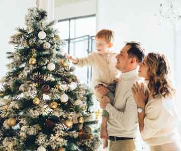 Family of 3 beside a Christmas tree, child holding an ornament.