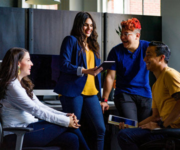 Four Happy Coworkers Hanging Out Inside The Office At Black Work Desks Near Plants And Looking At A Silver Tablet