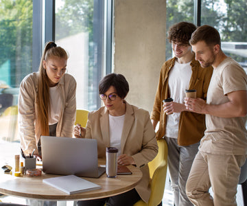 Four Coworkers In Bright Office With Large Windows Holding Morning Coffee And Working On Project Together At Table