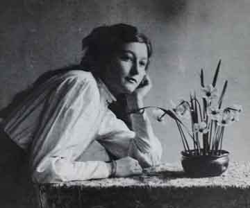 Black and white photo of woman at desk, hands on face, with small flower vase.