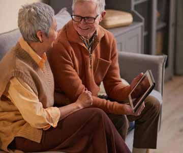 Smiling Old Man With Glasses Sitting Beside An Old Woman On Couch In Brightly Lit Living Room Near Dresser And Bookcase