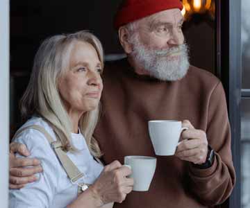 An older couple standing close and holding coffee cups, looking into the distance.
