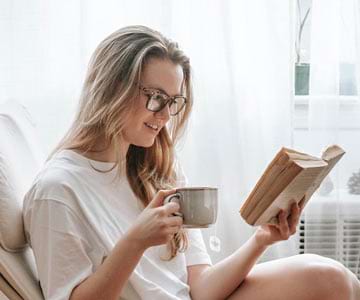 Happy woman reading a book and holding a cup of coffee near a window