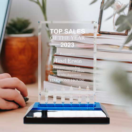 Custom Mirage Acrylic Award For Top Annual Sales With Logo And Name And Blue Mirror Base On Work Desk With Stack Of Books