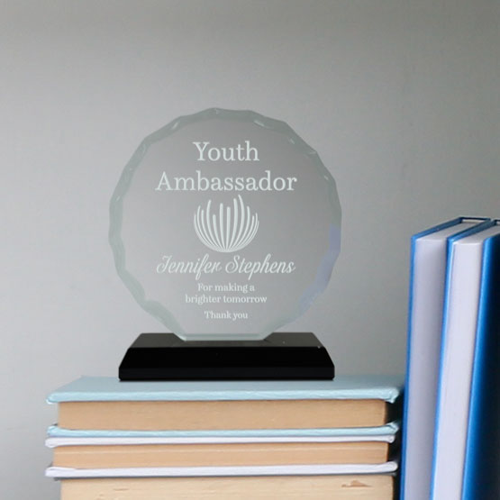 Custom Faceted Circle Glass Award For Youth Ambassador With Small Black Base Standing On Stack Of Blue Books