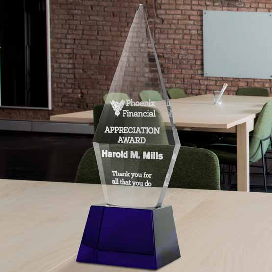 Custom Faceted Obelisk Glass Award For Appreciation With Blue Pedestal Base On Wood Work Table In Office With Brick Walls