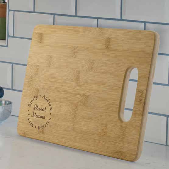 Custom Bamboo Cutting Board With Blessed Mama Text And Family Names Sitting On White Kitchen Countertop Against Backsplash