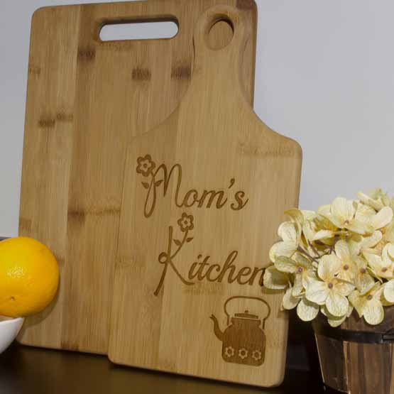 Custom Paddle Shaped Bamboo Cutting Board With Teapot Artwork And Mom’s Kitchen Text On Counter With Flower Pot