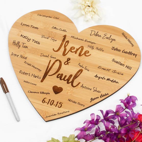 Custom Heart Shaped Bamboo Sign Engraved With Couple Names And Date And Marked With Guest Names On White Table With Flowers