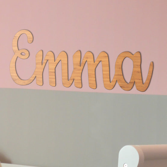 Custom Cutout Bamboo Sign With Emma Name Shape In Cursive Hanging On A Pink And Green Living Room Wall