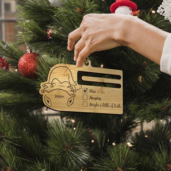Woman Hand Hanging Custom Bamboo Money Holder Ornament With Elf Hat Art And Naughty Or Nice Checklist Onto A Christmas Tree