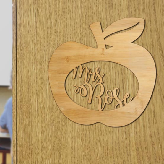 Custom Apple Shaped Bamboo Sign With Teacher Name Inside Oval Cutout Hanging On An Open Classroom Door