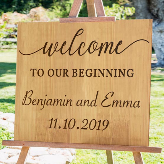 Custom Bamboo Sign Engraved With Couple Names And Date And Welcome To Our Beginning Text Standing Outside Via Wood Easel