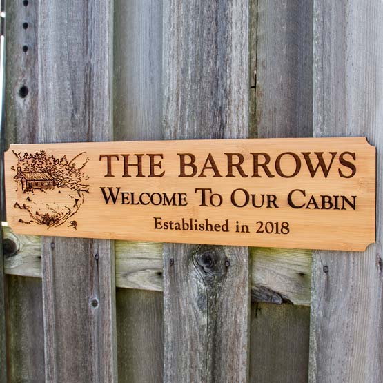 Custom Bamboo Sign Engraved With Cabin Art And Name And Date And Welcome To Our Cabin Text Hanging On Wood Fence Outside