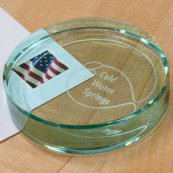 Custom Circle Glass Paperweight Engraved With Cold Water Springs Text And Logo Sitting On Office Desk With Stamped Envelope