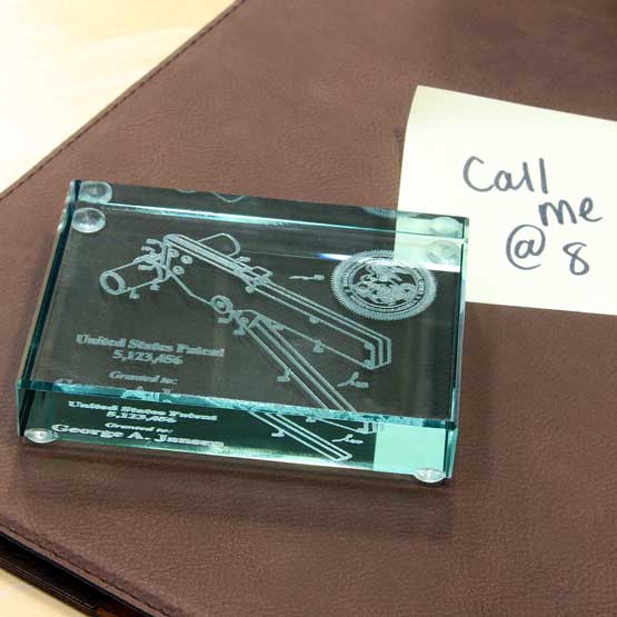 Custom Glass Paperweight Engraved With Patent Image And Details Sitting On A Leather Notebook With Post It Reminder
