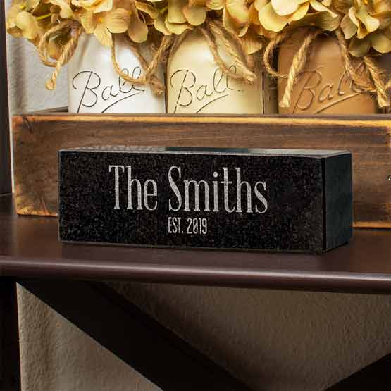 Custom Granite Pillar With Light Flecks And Engraved With Name And Date Sitting On Living Room Mantle Beside Flowers