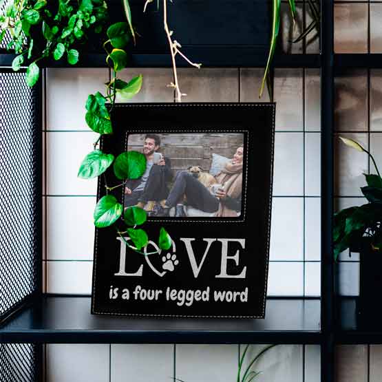 Black And Silver Faux Leather Picture Frame With Photo Of Couple And Dog And Love Engraving On Black Bookshelf With Plants