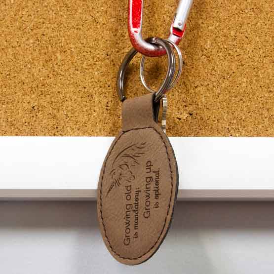 Dark Brown Faux Leather Oval Keychain Engraved With Unicorn Art And Growing Up Is Optional Message Hanging On Corkboard