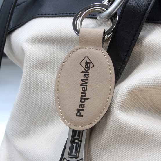Custom Light Brown Faux Leather Oval Keychain Laser Engraved With PlaqueMaker Logo Hanging From Beige And Black Handbag