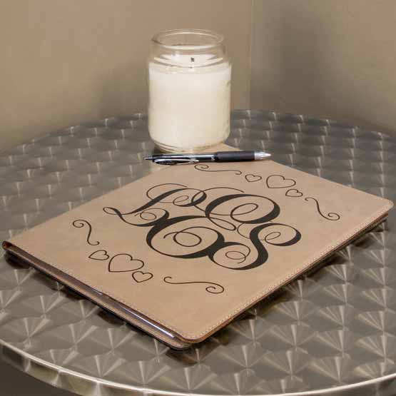 Light Brown Faux Leather Notebook Engraved With Ornate Initials And Heart Artwork On A Metal Side Table With Pen And Candle