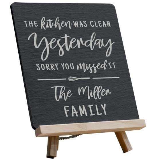 Custom Slate Stone Sign Printed With Fun Kitchen Message And Family Name Standing Via Wooden Easel Against White Background