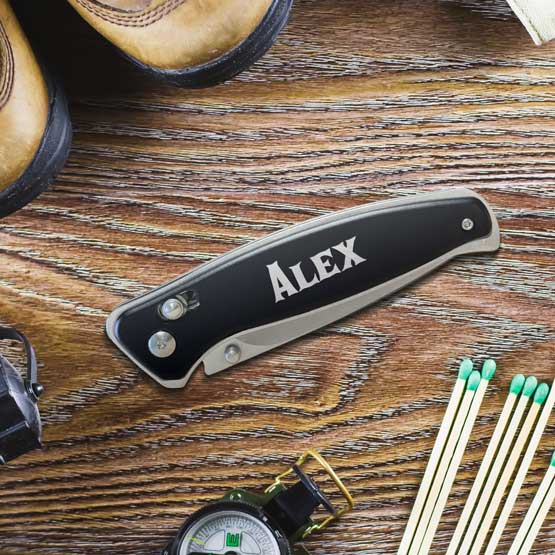 Custom Black Stainless Steel Folding Knife With Button Lock And Laser Engraved With Alex On Wood Table With Outdoorsy Tools