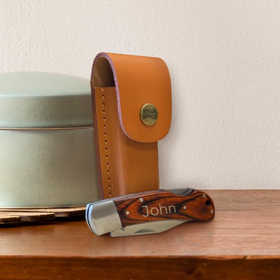 Custom Stainless Steel Folding Knife With Wood Handle Engraved With Name Beside Leather Sheath On A Bedroom Dresser