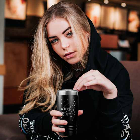 Young Woman On Chair Holding Black Stainless Steel Coffee Tumbler Engraved With Moon And Stars Art And Witch’s Brew Text