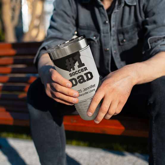 Man On Bench Holding Faux Leather Wrapped Coffee Tumbler With Soccer Theme And Engraved With Number One Dad Message