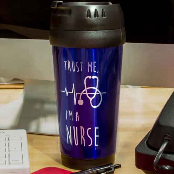 Blue Travel Mug With Lid And Engraved With Stethoscope Art And Trust Me I’m A Nurse Message On Busy Work Desk With Phone