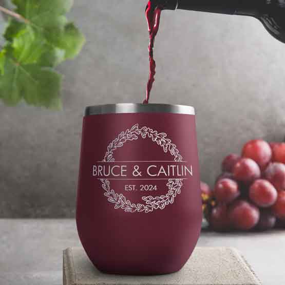Wine Bottle Pouring Wine Into Maroon Stemless Wine Tumbler Engraved With Couple Names And Wheat Art On A Kitchen Counter