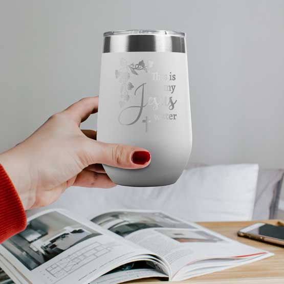 Woman Hand Holding White 16 Ounce Stemless Wine Tumbler Engraved With Grapevines And Cross And Jesus Water Message Above Desk
