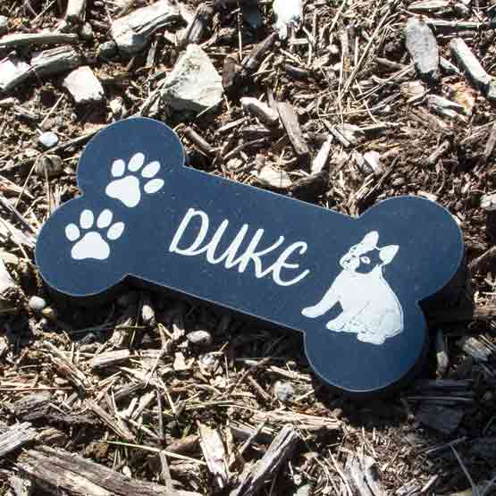 Bone Shaped Granite Dog Memorial Headstone Engraved With Name And Artwork Of Dog And Paw Prints Outdoors In Mulch