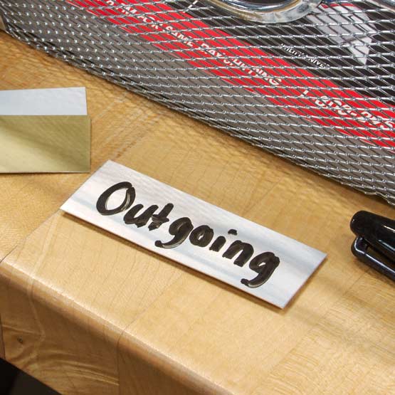 White Aluminum Tag Marked With Handwritten Outgoing Text On A Wooden Desk With Black Marker And Two Blank Tags
