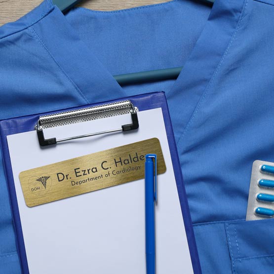 Custom Engraved Brass Medical Name Tag Laying On Clipboard And Scrubs. 