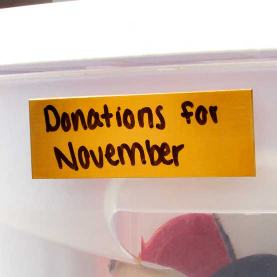 Bronze Name Tag Marked With Handwritten Donations For November Text In Black Fastened To A Clear Plastic Bin