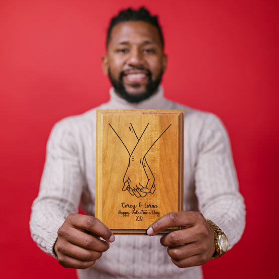 Man Holding Up Solid Cherry Plaque Engraved With Holding Hands Artwork And Names And Date And Happy Valentine’s Day Text