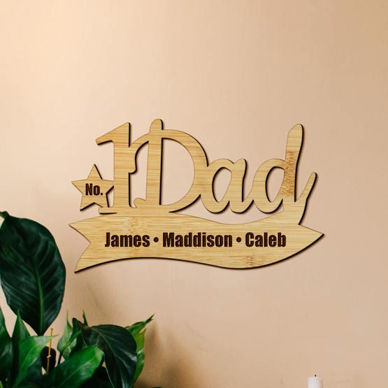 Bamboo Cutout Sign With Number One Dad Text And Star On Ribbon Shape Engraved With Names Mounted On Wall Above Plant