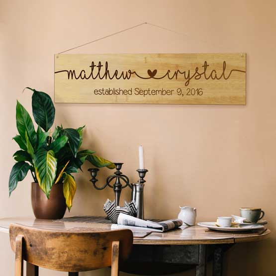 Rectangle Bamboo Sign Engraved With Cursive Couple Names And Established Date Mounted On Wall Above Dining Table Via Twine