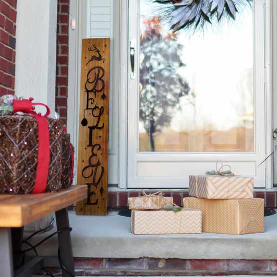 Bamboo Sign Laser Engraved With Believe Text And Reindeer Art Leaning Beside Front Porch Door Near Wrapped Presents