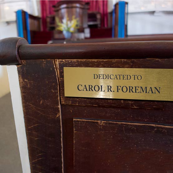 Custom Brass Dedication Sign Laser Fused With Dedicated To Carol R Foreman Message Mounted On Back Of Church Pew Indoors