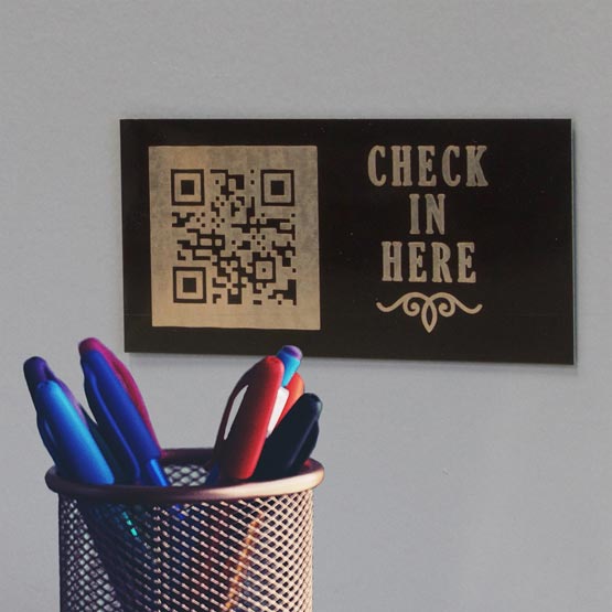 Black And Gold Aluminum QR Tag Laser Engraved With QR Code And Check In Here Text Mounted On Wall Above Cup Of Pens