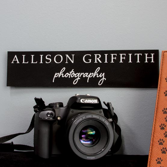 Black And Silver Faux Leather Sign Laser Engraved With Allison Griffith Photography Message On Wall Above Shelf With Camera