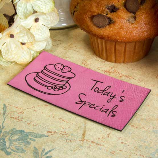 Pink And Black Faux Leather Label Engraved With Cake Art And Today’s Specials Text On Counter Beside Flowers And Muffin