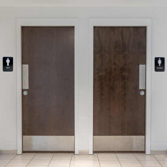 Two Public Restroom Doors With A Plastic Black And White Sign For Men And One For Women With Icons And Text And Braille
