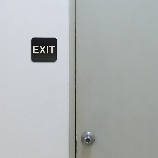Black And White Plastic Sign With Rounded Corners And Exit Text Above Corresponding Braille Mounted On Wall Beside Door